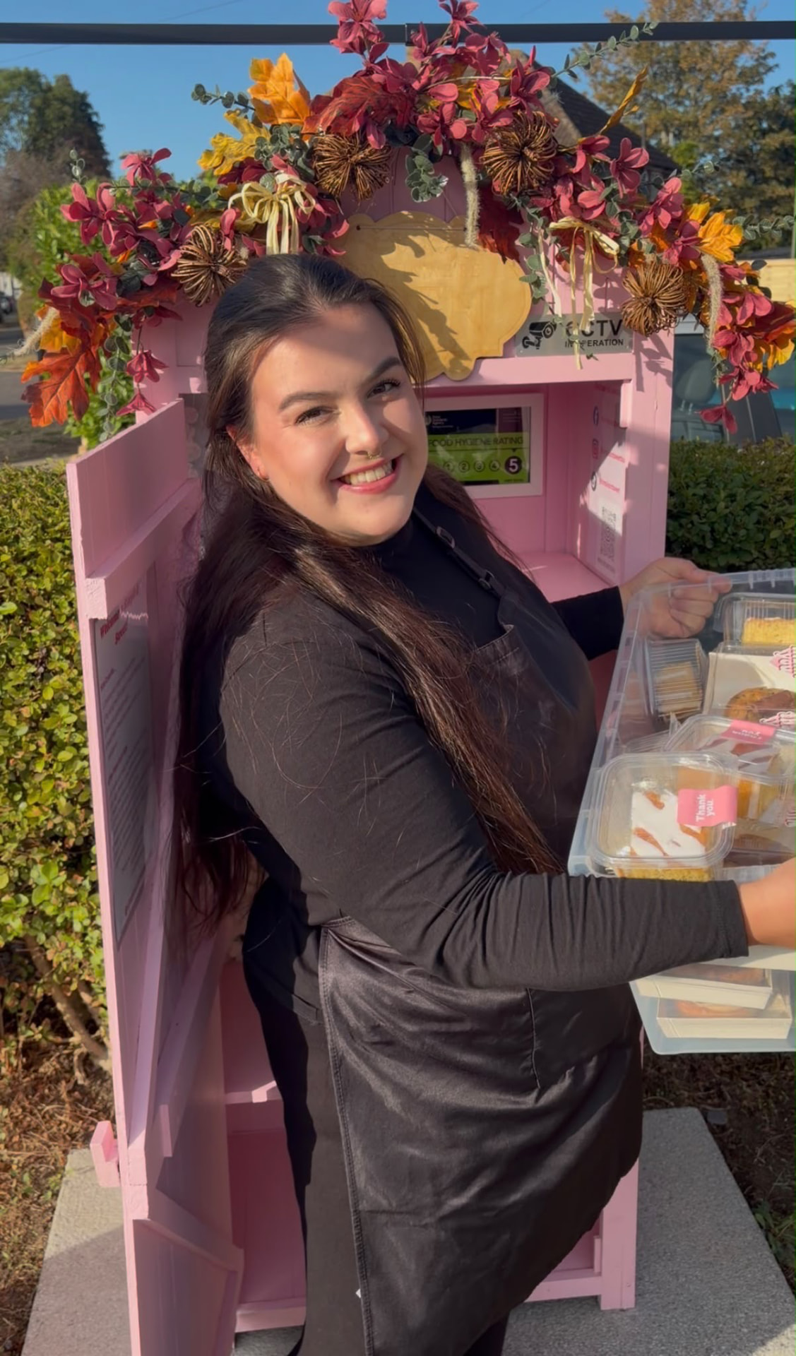 Billie smiling in a black apron while stocking the pink Cake Shed with trays of cakes